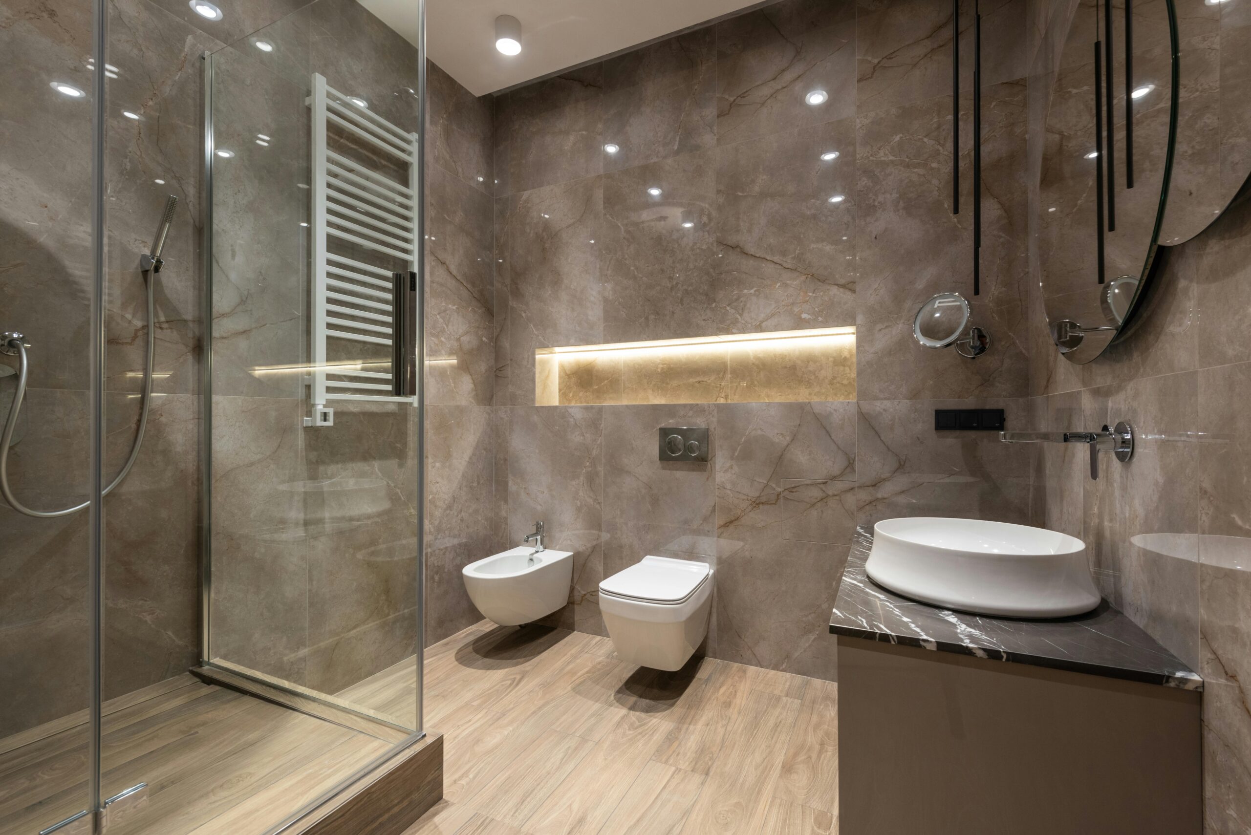 About - Beautiful, Stunning Bathroom Transformations by Expert Renovator Glass shower cabin and ceramic sink with toilet bowl and bidet in spacious contemporary bathroom with shiny tiles and round mirrors